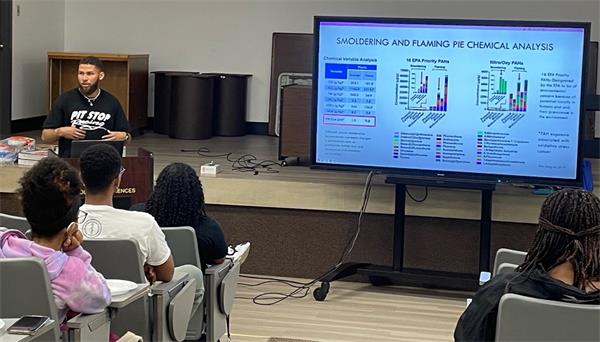 Keith Rogers is standing at the front of the room next to a big screen, talking to a crowd.